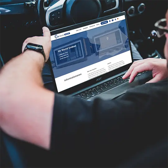 A guy sitting in the driver's seat of a car with a laptop on his lap. A website can be seen on the screen.