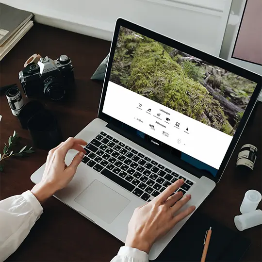 A laptop on a table, surrounded by camera equipment. A website can be seen on the screen.