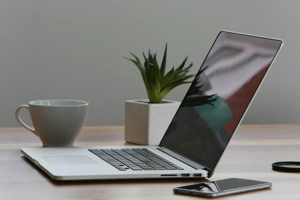 A beautifully arranged Macbook on a wooden table in front of a gray wall, on the table there is also an iphone, a plant in a pot and a cup.