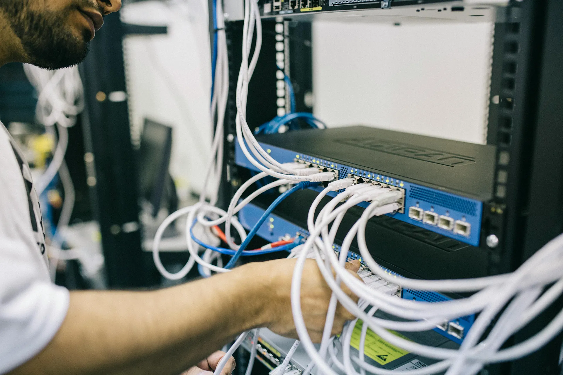 One guy plugs network cables into a switch located in a server rack. Other switches with network cables can be seen in this rack.