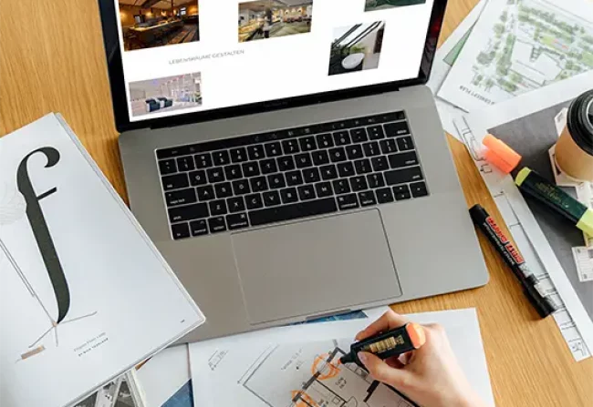 A laptop on a table, surrounded by plans. A website can be seen on the screen.